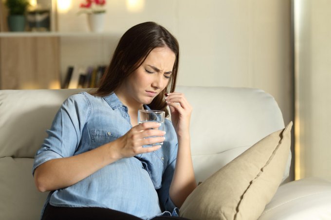 pregnant ill woman suffering and taking a pill sitting on a couch in the living room at home