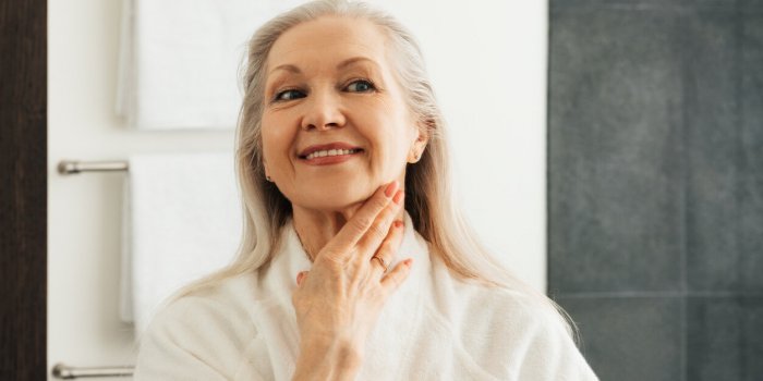 smiling senior woman touching skin on her neck in front of a mirror
