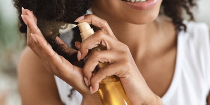 split ends repair treatment smiling african woman applying essential oil spray on her curly brown hair at home, cropped i...