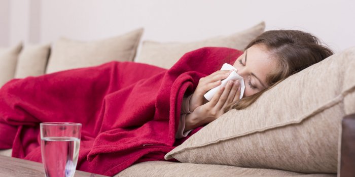 sick woman covered with a blanket lying in bed with high fever and a flu, blowing her nose pills and glass of water on th...