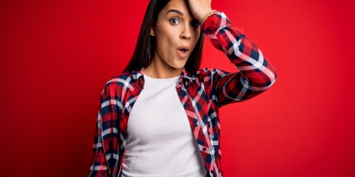 young beautiful brunette woman wearing casual shirt standing over isolated red background surprised with hand on head for...
