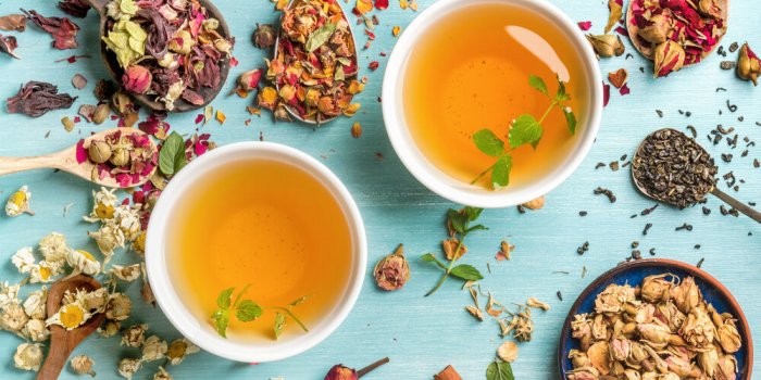 two cups of healthy herbal tea with mint, cinnamon ,dried rose and camomile flowers in spoons over blue background, top view