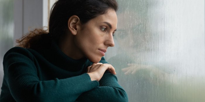 thoughtful stressed young hispanic latin woman sitting on windowsill, looking outside on rainy weather, having depressive...
