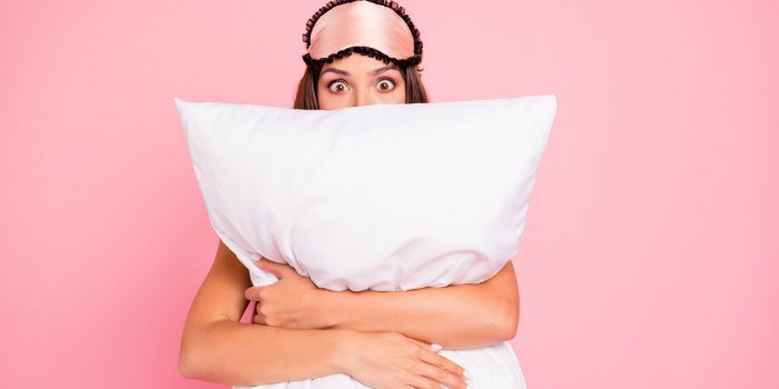 young gorgeous shocked lady wearing eye mask, hugging pillow, hiding isolated over pink pastel background