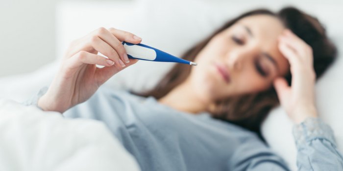 woman with flu virus lying in bed, she is measuring her temperature with a thermometer and touching her forehead