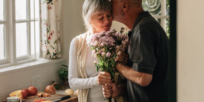 senior couple standing in kitchen holding a bunch of flowers senior man kissing his wife holding her hand at home
