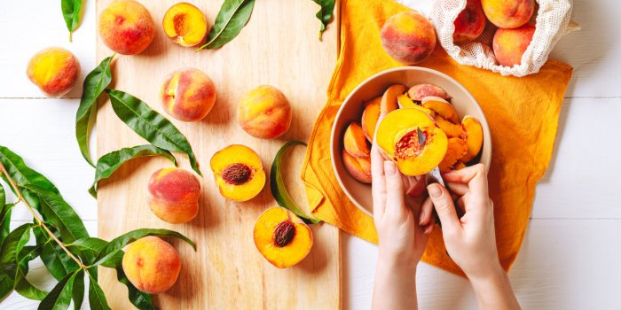 female hands cutting fresh sweet peaches peaches whole fruits leaves, half peach, peach slices on white wooden kitchen ta...