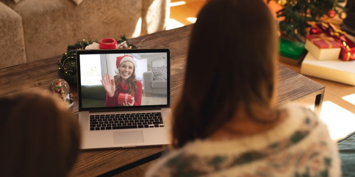 rear view of woman and son having a videocall with woman in santa hat waving while holding gift box on laptop at home soc...