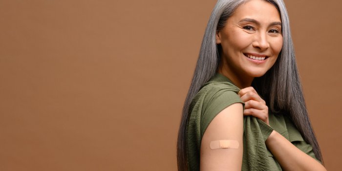smiling senior grey-haired woman with a medical patch after vaccination isolated on brown background, protecting hand wit...