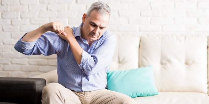 middle aged man sitting on the sofa and holding painful shoulder with another hand