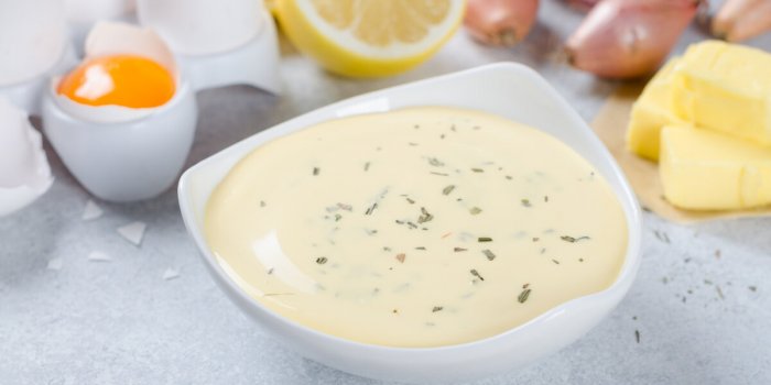 homemade basic french sauce bearnaise in a white bowl with ingredients, butter, shallot, lemon, eggs, on a light blue sto...