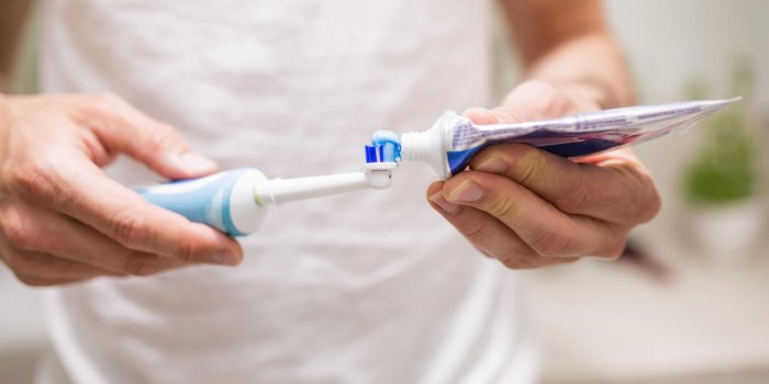 close up of human hands pouring blue toothpaste on an electric toothbrush man is blurred, in a white t-shirt there's bath...