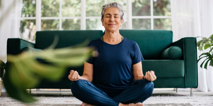 senior woman meditating in lotus position at home, sitting on the floor in fitness clothing mature woman doing a breathin...