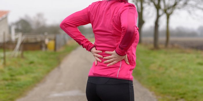 woman with back or kidney pain clutching her lower back as she takes a break from jogging or working out on a rural road ...