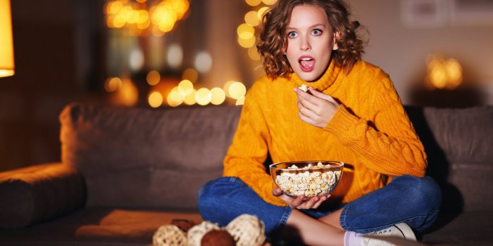 fascinated young female in sweater eating popcorn and watching interesting film on tv while sitting on couch in evening a...