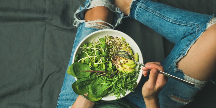 green vegan breakfast meal in bowl with spinach, arugula, avocado, seeds and sprouts girl in jeans holding fork with knee...