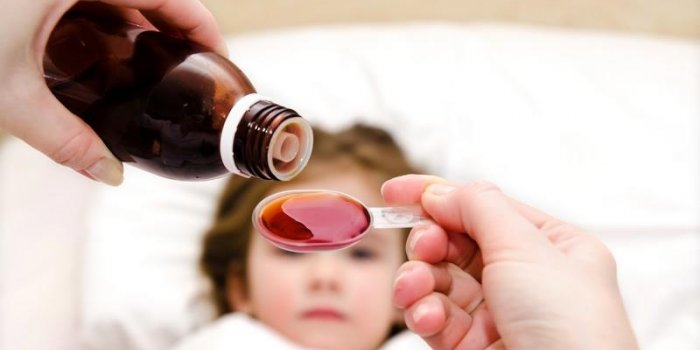mother pouring medication in a spoon in the foreground and sick little girl in the bed