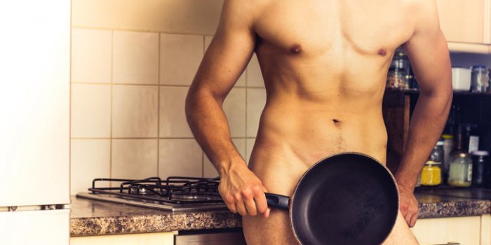 jeune homme nu avec les muscles tonique est debout devant la cuisinière dans une cuisine domestique couvrant ses parties...