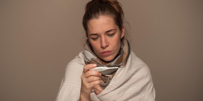 portrait of young woman looking on thermometer and wrapped in a blanket during illness on a gray background