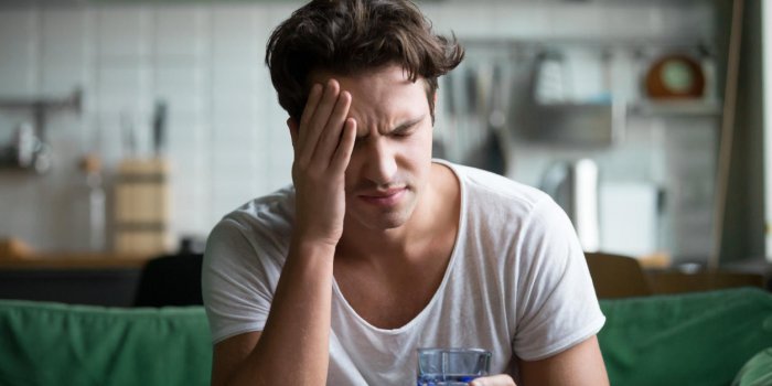 young man suffering from strong headache or migraine sitting with glass of water in the kitchen, millennial guy feeling i...