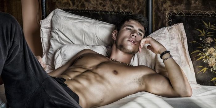 modèle masculin sexy torse nu couché seul sur son lit dans sa chambre, en détournant les yeux avec une attitude sédui...