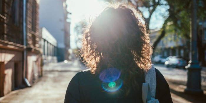 rear view young woman walking down the street, sun flare towards her female with curly hair goes along city sidewalk