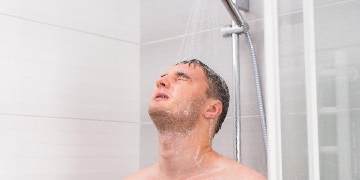young man with closed eyes taking a shower, standing under flowing water in shower cabin with transparent glass doors in ...