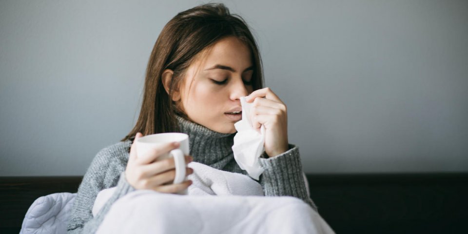 the attractive young woman is seen in her bed, feeling sick while blowing her nose and holding a mug