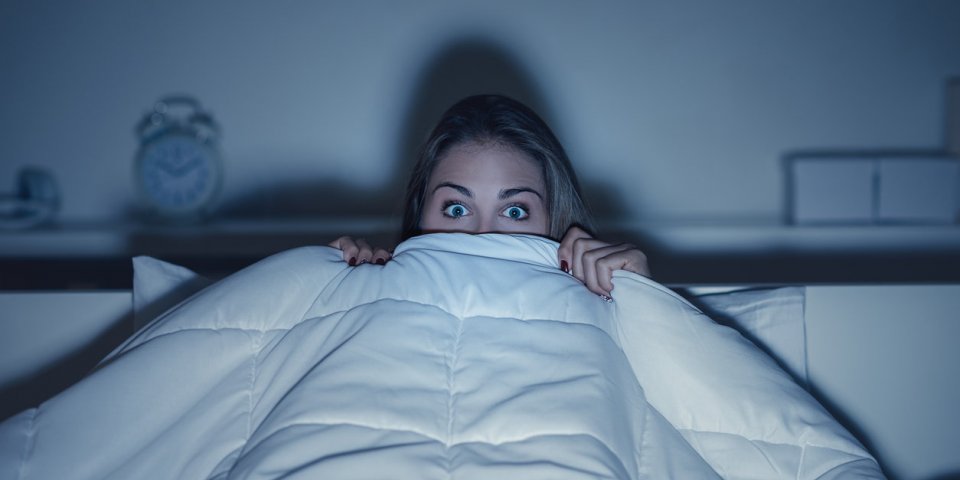 woman watching a scary horror movie on tv late at night, she is frightened and hiding under the blanket