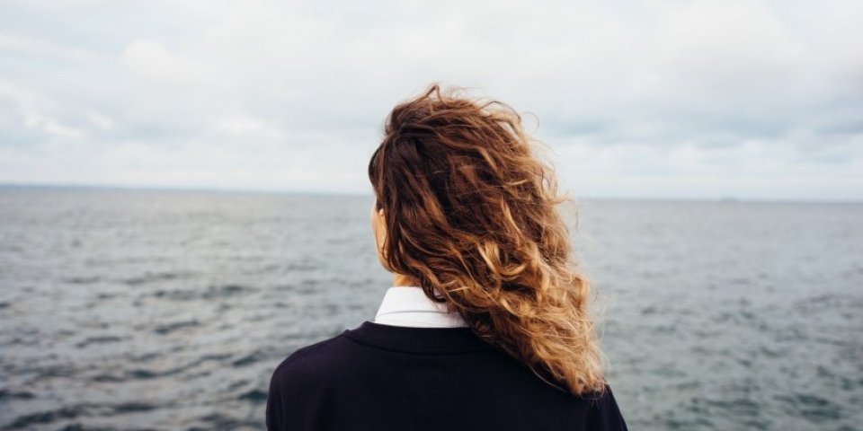 rear view of young woman looking at overcast sky and gray sea female with red curly hair standing alone thinking at the b...