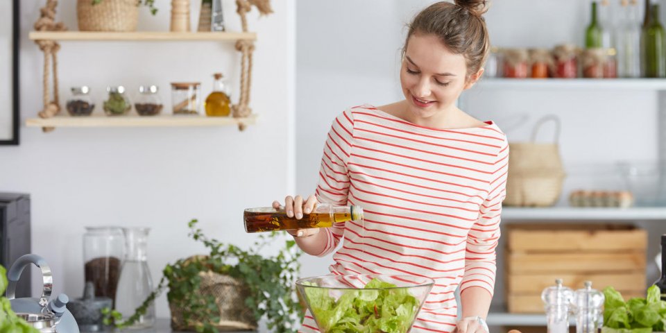 young woman standing in a kitchen and adding olive to vegetable salad