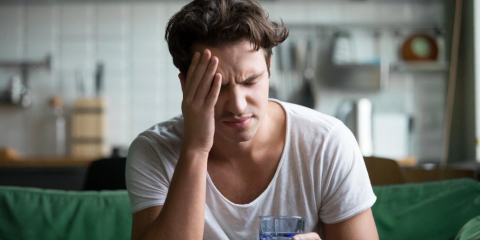 young man suffering from strong headache or migraine sitting with glass of water in the kitchen, millennial guy feeling i...