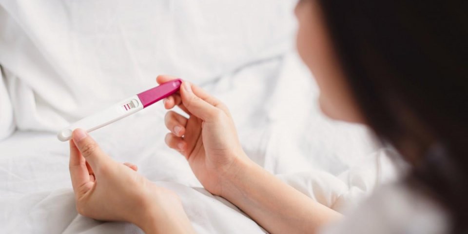 back view of happy smile woman looking at pregnancy test with positive result in hand on the bed