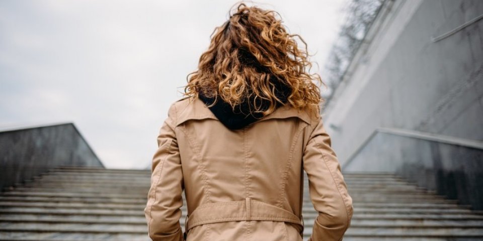 back view young woman with curly hair dressed in beige coat stands at bottom of marble stairs looking up