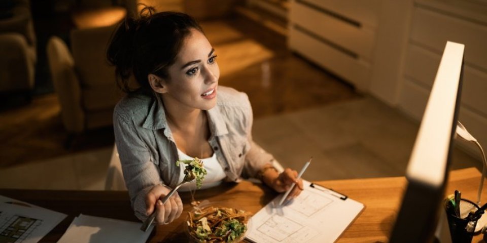 high angle view of young woman eating healthy salad while working on desktop pc in the evening at home