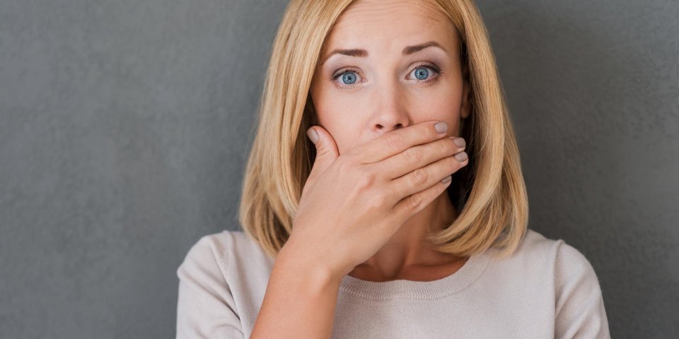 surprised mature woman covering mouth with hand and staring at camera while standing against grey background