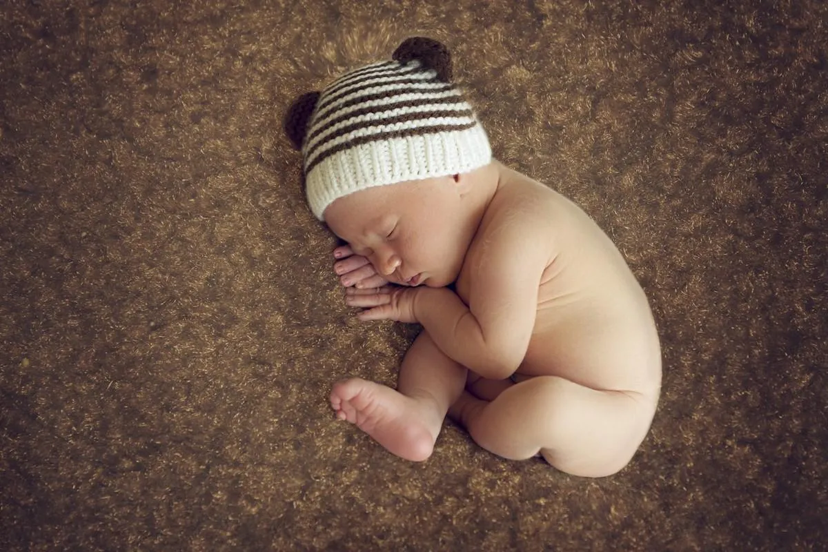newborn baby boy sleeping peacefully in the fetal position on a brown background with the beanie with ears in the image of teddies