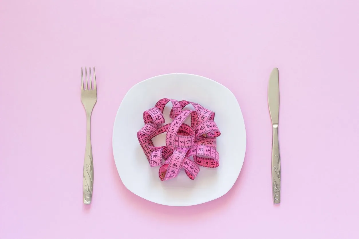pink measuring tape lying on plate in the form of spaghetti, knife and fork on pink background diet or anorexia concept top view