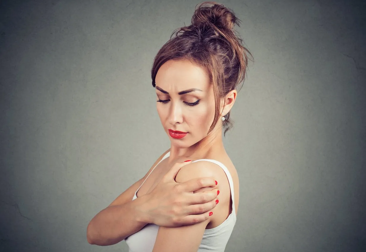 young brunette rubbing shoulder having pain in joint and standing on gray background