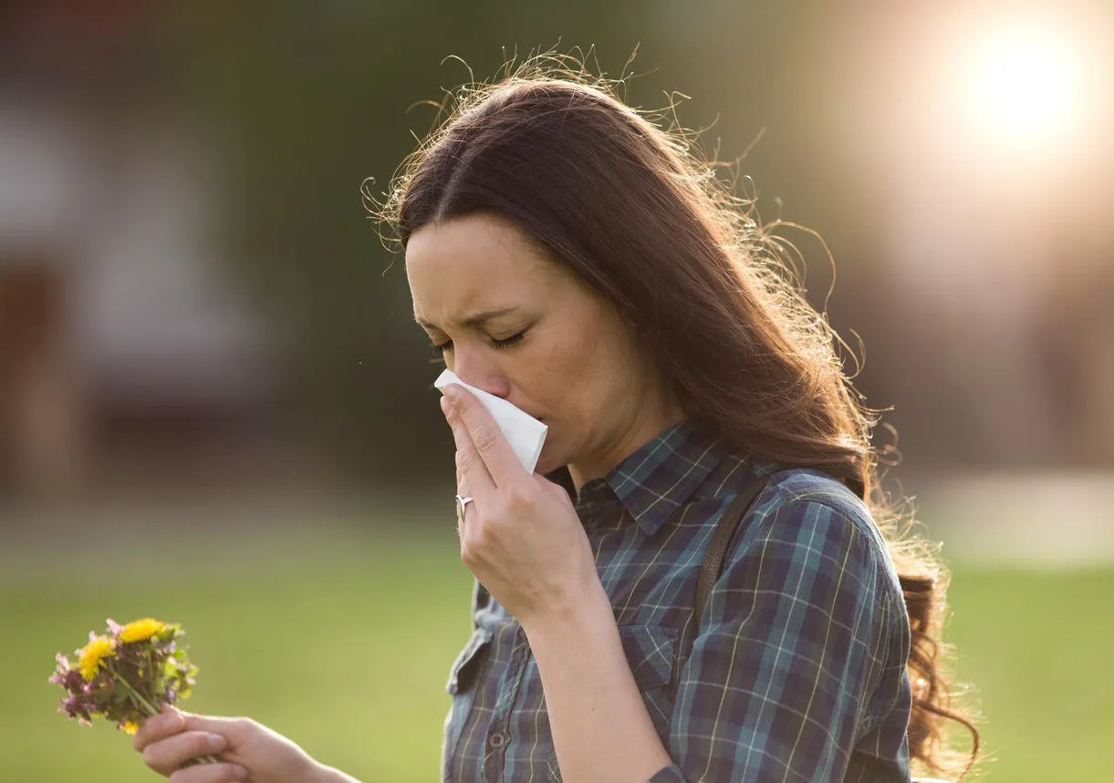 young pretty woman sneezing and blowing nose while holding bouquet of spring flowers from meadow pollen allergy symptoms