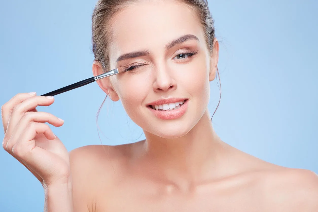 beauty concept, head and shoulders of young woman with closed eye, smiling closeup of girl looking at camera and touching eyelid with brush, painting with eyeshadows