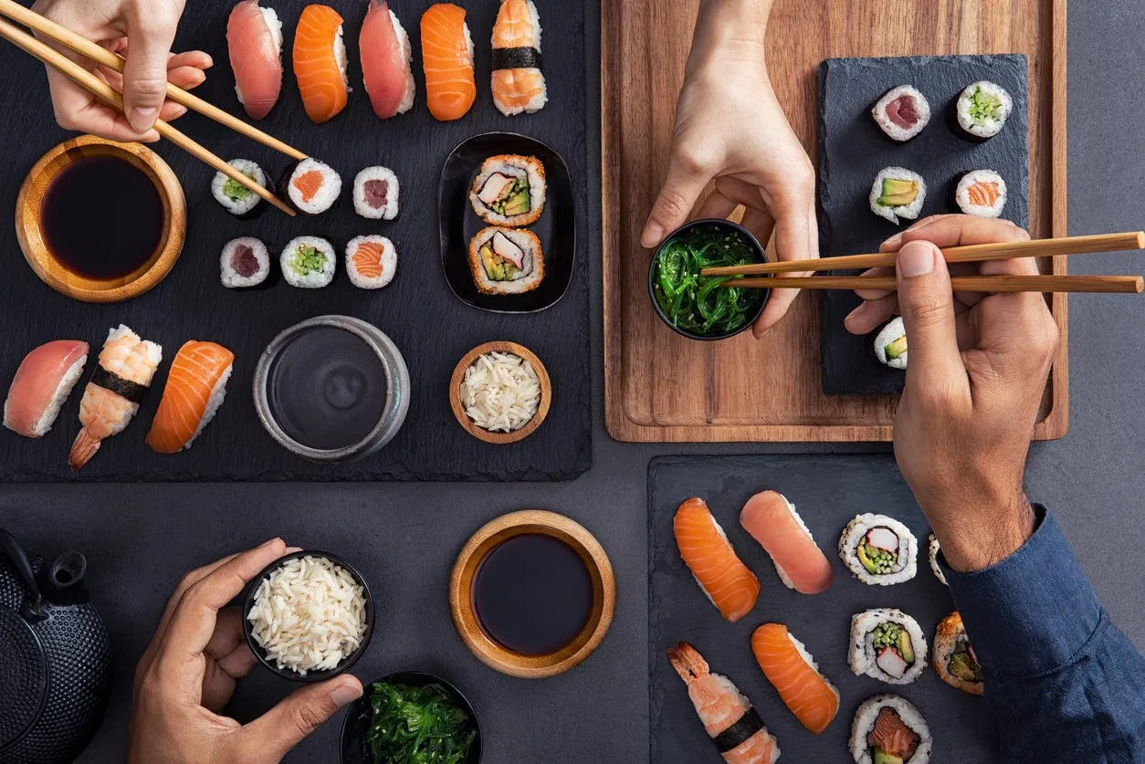 top view of couple hands eating sushi food at japanese restaurant high angle view of woman hand serving seaweed in little bowl with sesame to man while holding hosomaki with chopsticks couple eating and sharing sushi roll, maki, nigiri, uramaki