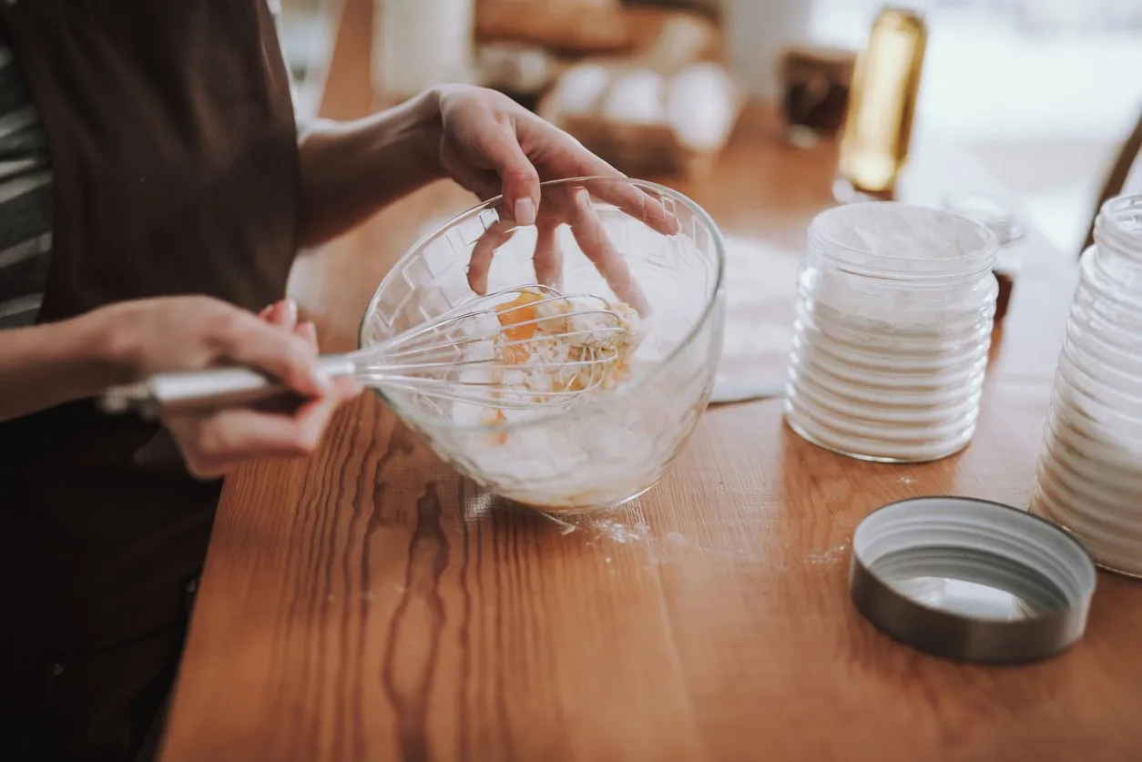 top view of female mixing flour with eggs while using whisk she is cooking bakery in domestic atmosphere