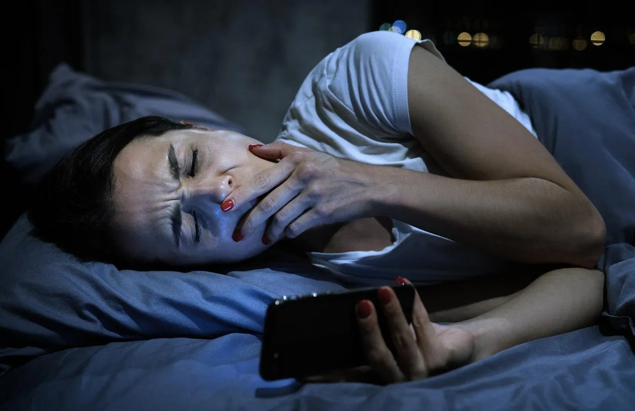 young sleepy female using mobile phone, yawning late at night, lying in bed smartphone addiction concept
