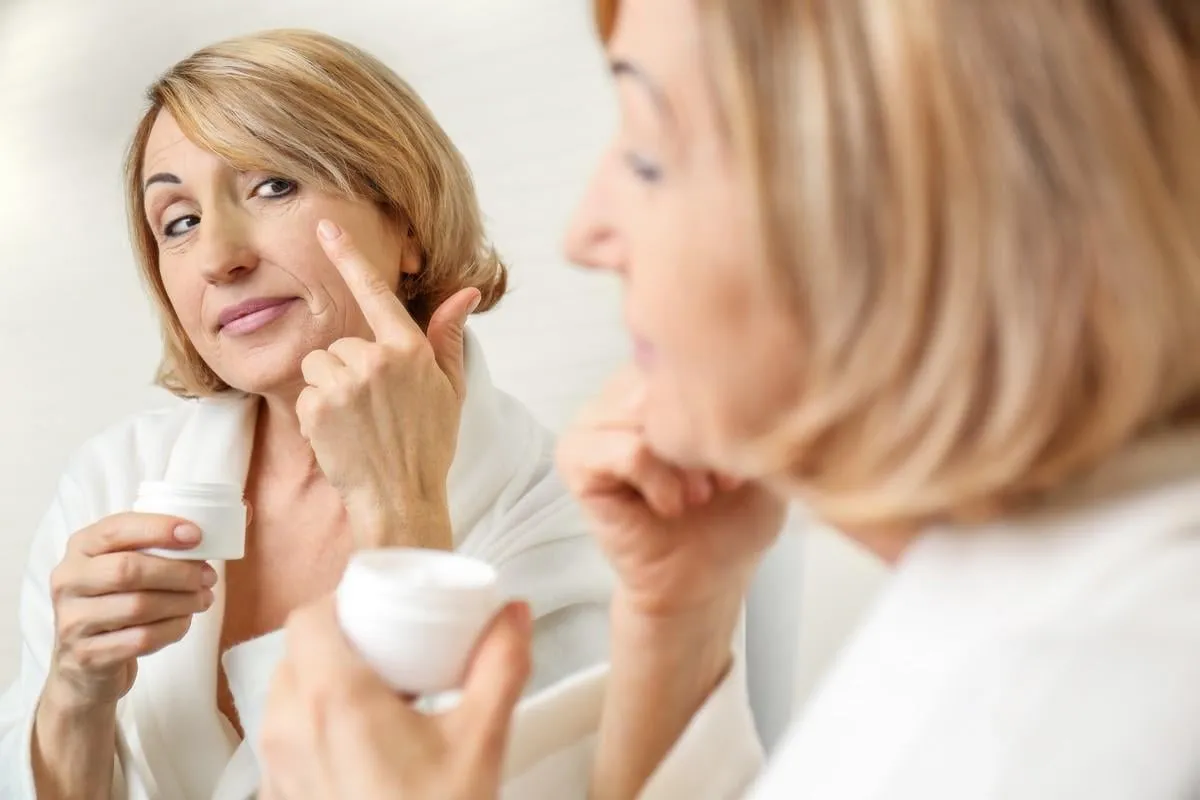 senior woman applying cosmetic cream on face near mirror