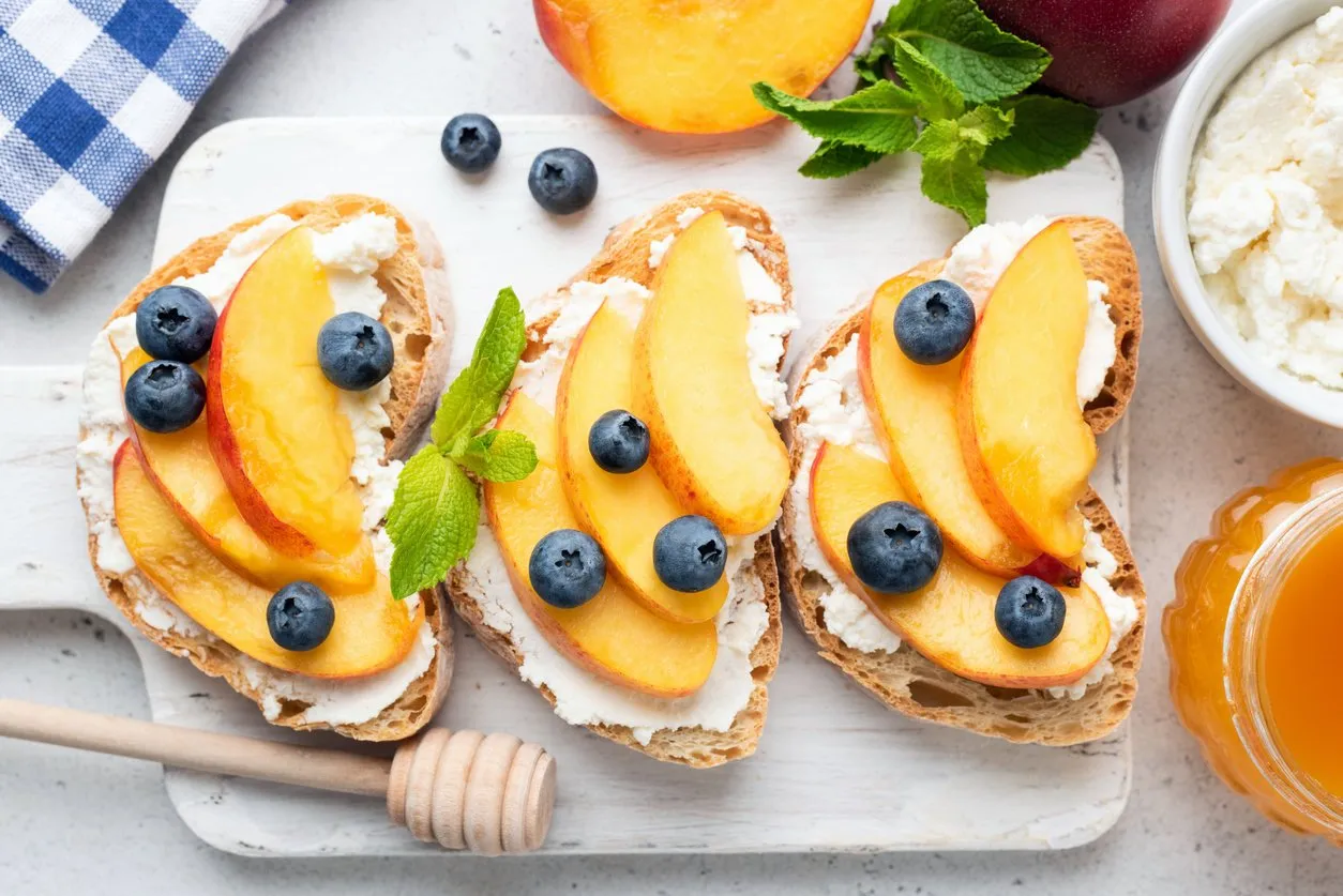 delicious toast with soft ricotta cheese, fresh juicy peach, blueberry and honey on white wooden board top view healthy summer appetizer or snack