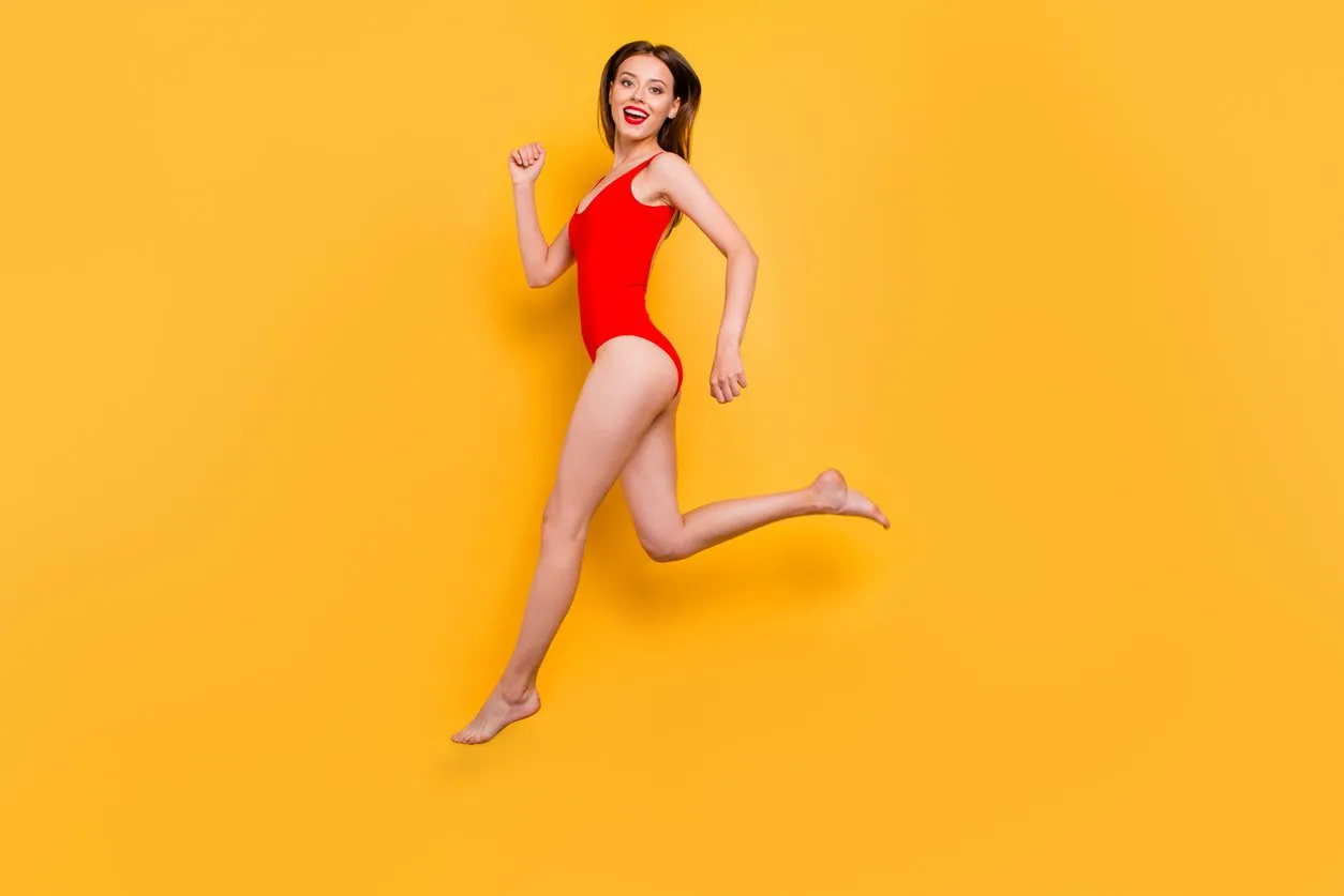 run to meet the summer full-legs portrait of fit sporty girl in red swimsuit jumping over in the air looking at camera isolated on yellow background
