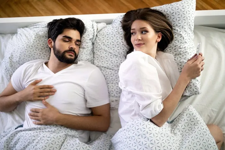 beautiful young wife texting with lover on smartphone while husband is sleeping