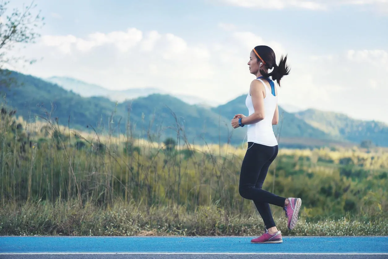 asian young fitness woman with smartwatch in white suit black trouser pink shoe running at blue street road green grass field and mountain background
