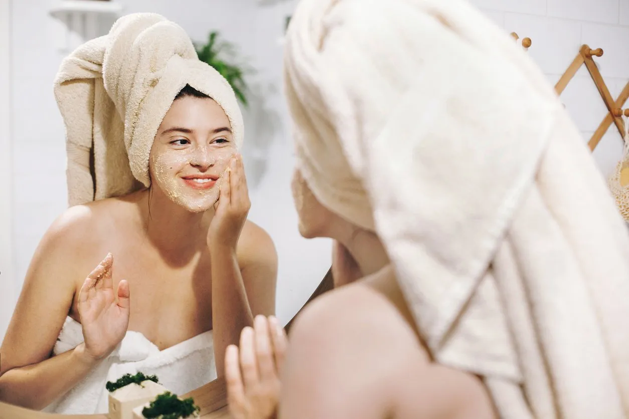 skin care concept young happy woman in towel making facial massage with organic face scrub and looking at mirror in stylish bathroom girl applying scrub cream, peeling and cleaning skin